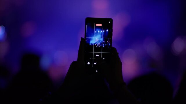 Hand holding smartphone with camera app open recording a live concert, purple bokeh background, stage performer visible on screen