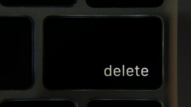 Close view of a finger pressing the Delete key on a laptop keyboard in a dark workspace as the backlight activates the symbol on the key. User input, command action.