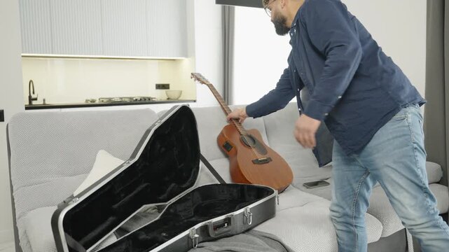 Man packing an acoustic guitar into a case while standing next to a sofa at home