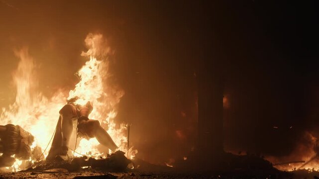 Slow motion. A falla figure silhouette stands engulfed in intense flames during La Crema in Valencia, ember sparks rising in the dark