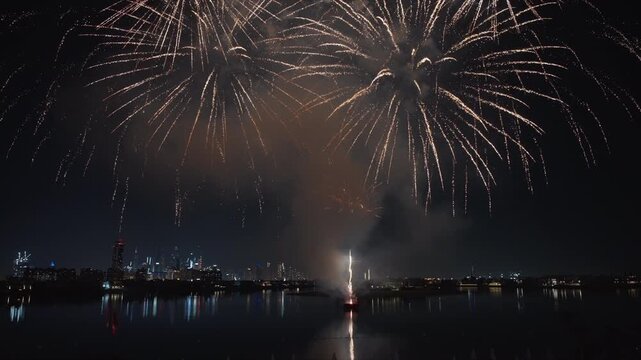 Spectacular golden fireworks illuminate the dark night sky above a glowing urban skyline with modern skyscrapers reflecting on calm harbor waters during a festive celebration event.