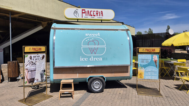 Blue ice cream trailer "Piaceria Sweet Ice Dream" with menu boards and yellow seating under a bright blue sky. Picture taken on April 27, 2026 in Bucharest, Romania.
