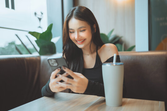 Beautiful young Asian woman smiling while using a smartphone at a modern cafe. She sits with a reusable tumbler, enjoying a relaxed indoor lifestyle and mobile connectivity. Perfect for tech, sustaina