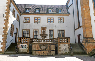 A quiet square with a central fountain lies in front of the symmetrical architecture of a historic monastery building, combining baroque elements with clean structural lines. © Heidi