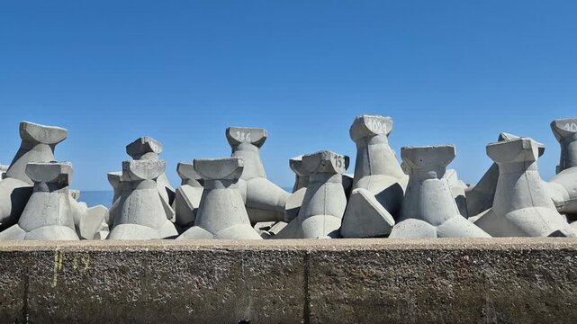 A row of gray concrete tetrapod breakwaters with identification numbers stands against a clear blue sky, designed to protect the coastline from erosion.