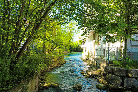 A lively river flows through a green urban landscape, framed by trees and accompanied by residential buildings along the riverbank in warm sunlight.