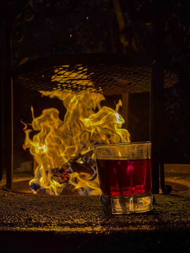 Warsaw, Poland, May 03, 2026. A glass of dark red liquid stands on a table next to a blazing fire at night. High quality photograph.