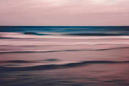 Long exposure captures gentle ocean waves as smooth pastel streaks beneath a calm horizon at dusk.