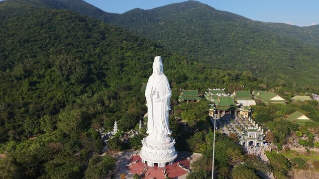 Vietnam, Da Nang Giant White Statue Towering Over Green Hills In Danang, Panoramic Aerial Sweep Reveals Temple Complex, Visitors At Base, Sunlit Serenity And Lush Forest Backdrop Creating