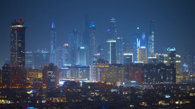 Stunning panoramic view of Dubai Marina skyline at night featuring illuminated skyscrapers rising through hazy atmosphere with residential neighborhood in foreground and deep blue twilight sky above.