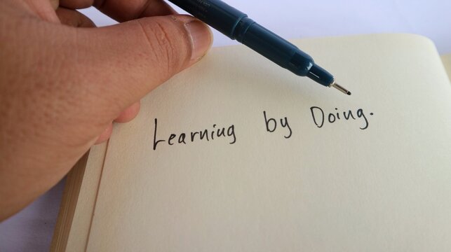 A text that says "learning by doing" written on a notebook with a pen on it.