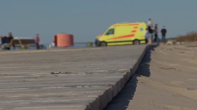 Emergency responders near sandy coast. Ambulance units advance along wooden pathway. Emergency responders march along wooden walkway with blurred vehicle background under warm sunlight