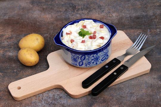 Hearty potato salad with bacon in a blue and white ceramic bowl on a wooden board.