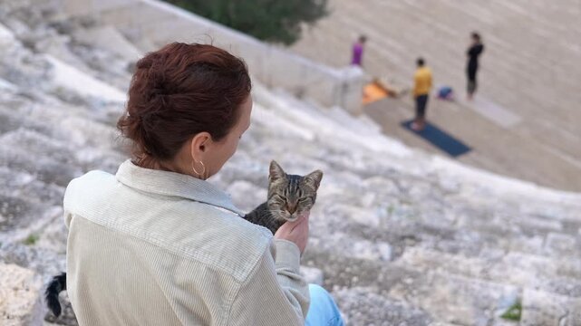 Woman petting a cat while people are practicing yoga. Woman quietly sitting outdoors, gently caressing cat's head while yoga students train softly in blurred background