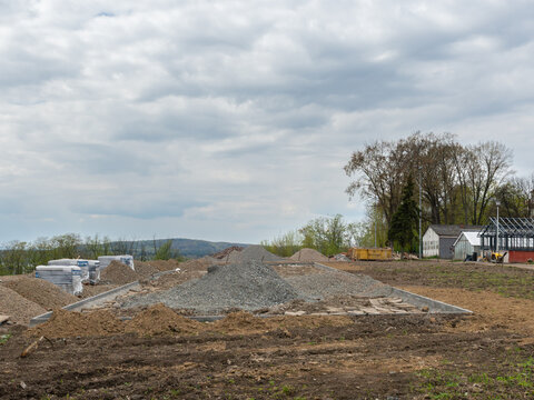 Building site with piles of gravel, sand and prepared concrete foundation, construction materials and equipment visible under cloudy sky, showing early stage residential development.