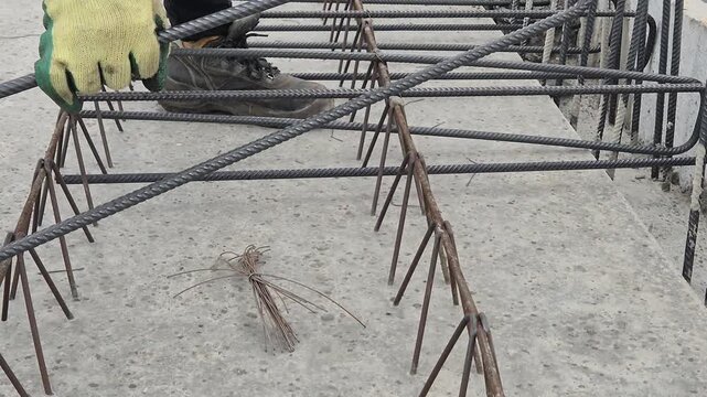 Slow motion side view of a construction worker's hands tying a steel stirrup with a hook tool and binding wire.