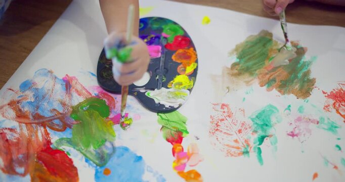 Children's hands use brushes and a palette to paint on paper, an educational activity focused on experiential learning and creative development in early childhood.