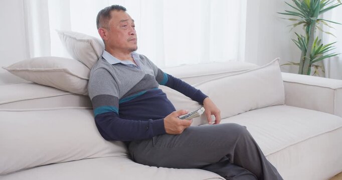 A senior man feeling bored sits on the couch, channel surfing with a remote control while unwinding alone in his bright living room during his retirement.