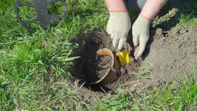 A treasure hunter wearing gloves and casual clothes digs a hole with a small yellow shovel, finding a clay pot or treasure, while searching for buried objects in a grassy open space