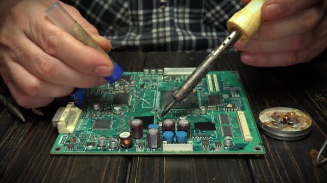 Hands working on a circuit board with soldering iron and tools, applying solder and flux in a well-lit workspace with wooden table surface