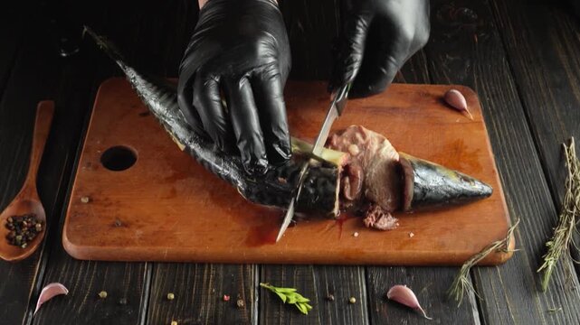 Chef in black gloves skillfully fillets a mackerel fish on a wooden cutting board with garlic cloves and herbs arranged around the scene
