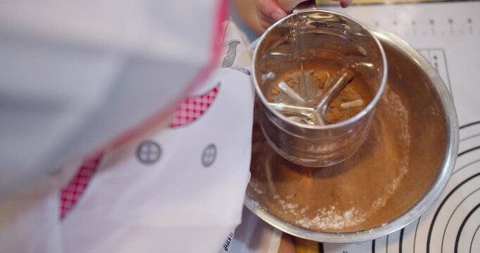 A baker carefully sifts cocoa powder with a metal sieve into a bowl, preparing the dry ingredients for a homemade chocolate dessert recipe from a high angle view.