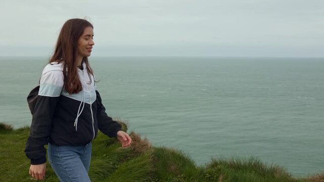 Happy young female tourist with a backpack walking along the edge of the rocky cliffs of &Eacute;tretat, France, enjoying the ocean view, wind blowing through her hair, peaceful solo travel moment, feeling f