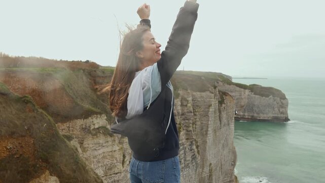Young woman standing on the edge of the cliffs of tretat, France, raising her arms in triumph, symbolizing success, achievement and dreams coming true, feeling absolute happiness and inspiration, cine