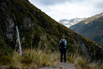 Fototapeta premium Hiker Standing on Mountain Trail Admiring Waterfall in New Zealand's Southern Alps