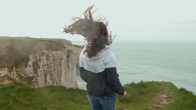 Young woman reaching the cliffs of &Eacute;tretat, France, joyfully jumping in place and raising her arms, celebrating arrival, admiring breathtaking ocean view, feeling happiness, freedom and excitement, ci