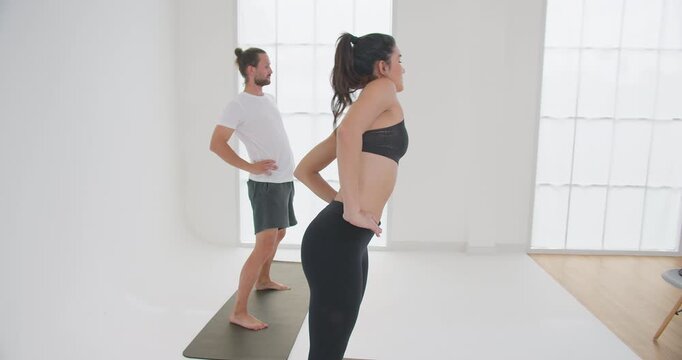 A young woman and man practice a standing backbend exercise in a bright minimalist studio, focusing on core strength and improving their physical wellbeing.