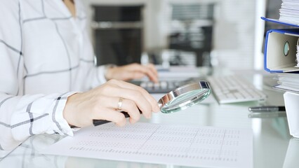 Fototapeta premium Businesswoman hands analyzing financial document with magnifying glass, working on computer in office, reviewing data. Audit and taxes in business