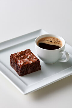 Overhead view of chocolate brownie with sea salt and coffee on a white plate against a clean white background.