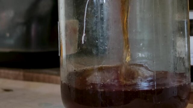 Kombucha is prepared in a large glass jar during the fermentation process. The drink is then poured into a glass, showing a natural homemade beverage in a clean and simple setting. 