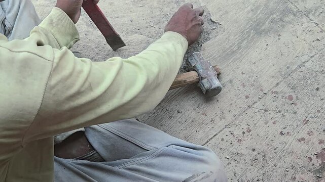 A laborer uses a hammer and chisel to break a concrete slab. This manual technique is essential for precision in waterproofing, floor repairs, and artisanal stonework on traditional construction sites