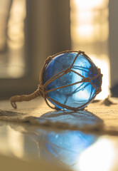 Vertical composition of blue glass fishing float with rope net and reflection in warm indoor light setting. © Trygve