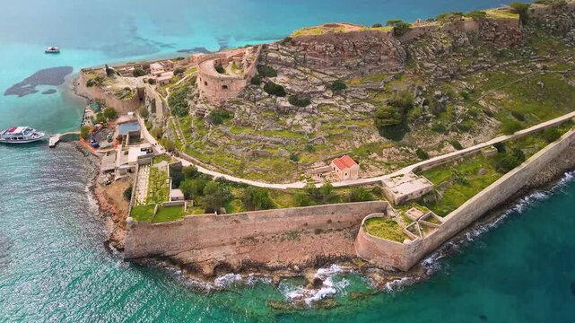 Aerial close-up flight over Spinalonga fortress with tourist boat in Crete, Greece. Drone view of Venetian castle ruins, turquoise sea and ancient stone walls from above. Scenic travel video, 4k