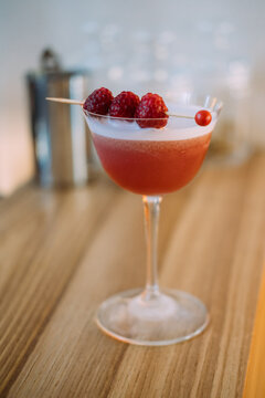 Served cocktail. Pub beverage. Red drink with frothy white top in stemmed glass. Refreshing mix with raspberry aroma flavor on wooden counter.