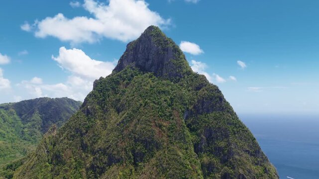 Drone footage captures the majestic Piton volcanic peak covered in lush green tropical rainforest on Saint Lucia island. Dramatic cliffs meet the blue Caribbean Sea under sunny skies with white.