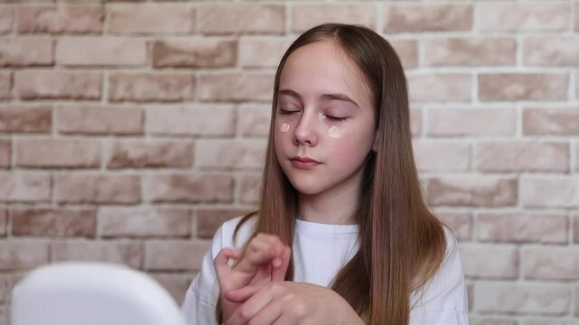 A pretty young blonde girl applies concealer on her face while sitting in front of a mirror. The Alpha Gene. Teen skin care