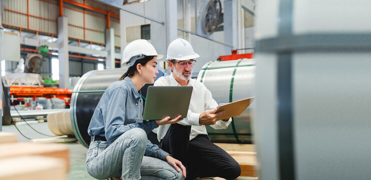 Two Caucasian men are collaboratively reviewing technical documents and inspecting metal sheets in brightly lit industrial factory setting, demonstrating focused quality control and professional