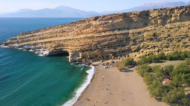 Aerial view of Matala beach at golden hour, Crete, Greece. Drone panning shot from ancient rock caves to coastal village with golden sunset light on sand and turquoise sea. Scenic landscape, 4k