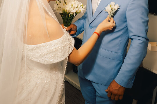 Couple prepares for wedding ceremony with flowers and attire in  venue setting.woman in a wedding dress places  flower on  man's suit while holding  bouquet.  ready for wedding in  venue.