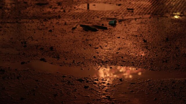 Wet muddy urban ground at night with tire tracks and scattered debris, shallow puddle reflecting warm orange and amber fire light