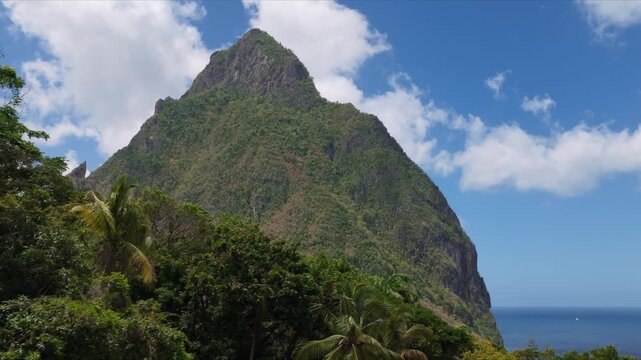 Majestic Piton volcanic peak rises dramatically against bright blue sky with scattered white clouds, lush tropical rainforest covering steep slopes while calm Caribbean Sea stretches to the horizon.