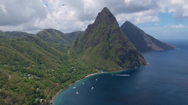 Aerial footage of iconic Piton volcanic peaks towering over lush tropical rainforest and turquoise Caribbean Sea in Saint Lucia. Sailboats anchor in calm bay waters below dramatic twin mountains.