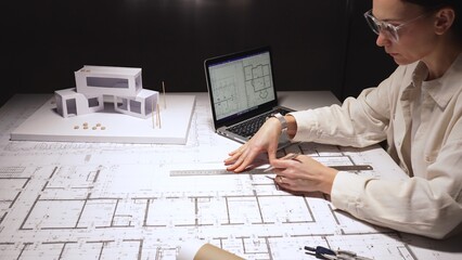 Architect drawing architectural floor plans on a large blueprint paper, working on a complex residential project with a laptop displaying cad designs and a physical house model on the desk © rogerphoto