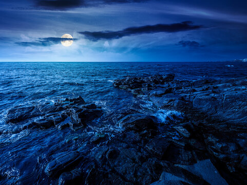 sea coast with stones and boulders at night. view of beach with rock in seaweed. full moon over dynamic cloudscape on the sky above horizon. mysterious summer vacations background concept