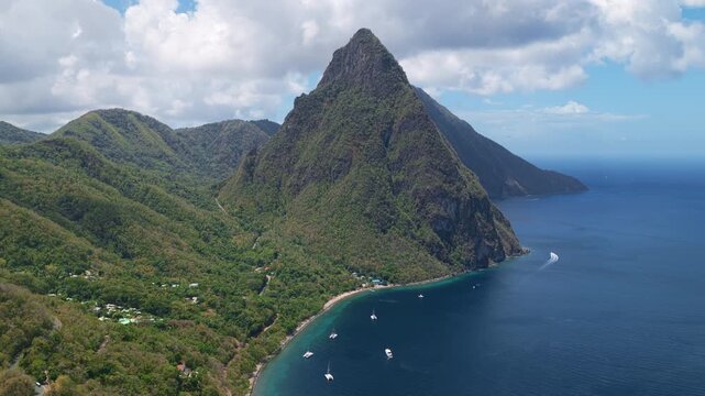 Aerial drone footage of the iconic Piton volcanic peaks in Saint Lucia with lush green rainforest slopes meeting turquoise Caribbean waters. Boats anchor in the bay below dramatic cliffs.