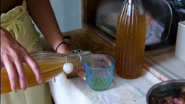 Kombucha is prepared in a large glass jar during the fermentation process. The drink is then poured into a glass, showing a natural homemade beverage in a clean and simple setting. 